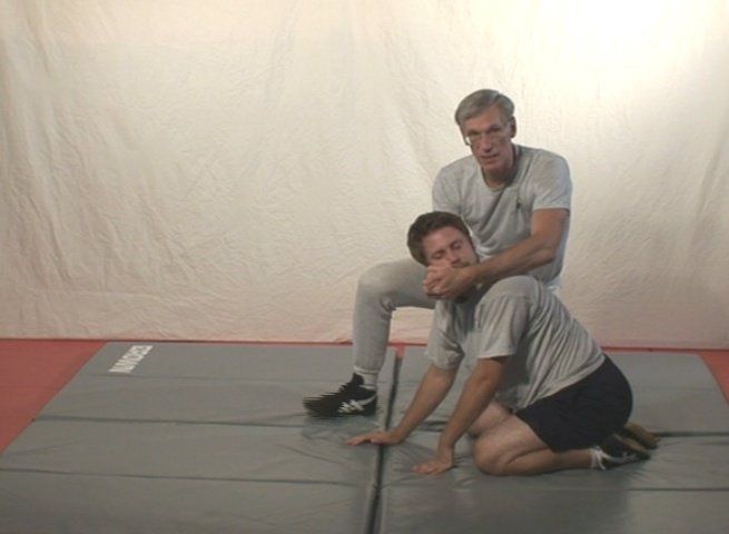 Man demonstrating a chokehold on another person kneeling on a mat. One person behind, one person on knees.