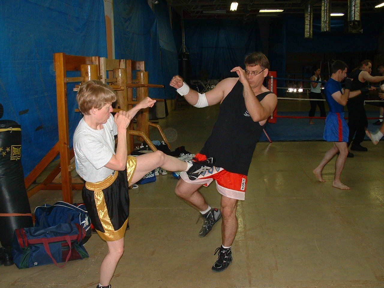 Two people sparring in a gym, one kicking. Others practice in the background.