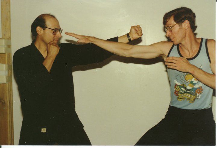 Two men practice martial arts with sticks. One blocks while the other attempts a punch. Indoor setting.