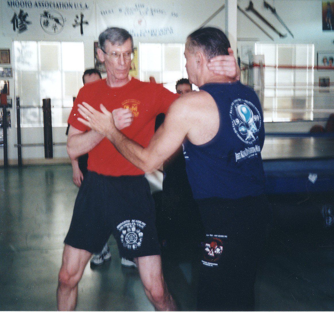 Two men in a gym practice a self-defense move. One in red shirt blocks, the other in blue, grips his neck.