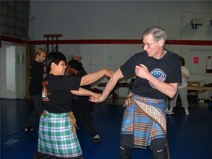 Two people practicing martial arts in a gym. Both wearing matching tartan skirts and t-shirts, one blocking a punch.
