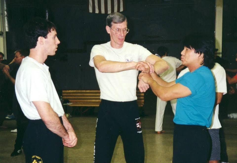 Two people practicing martial arts in a gym, each wearing a patterned wrap and black t-shirts. Others watch in the background.