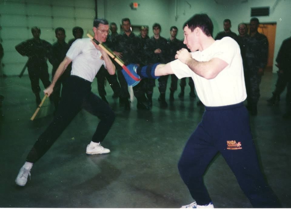 Two people practice martial arts, one with a stick, the other punches at a padded target. Soldiers watch.
