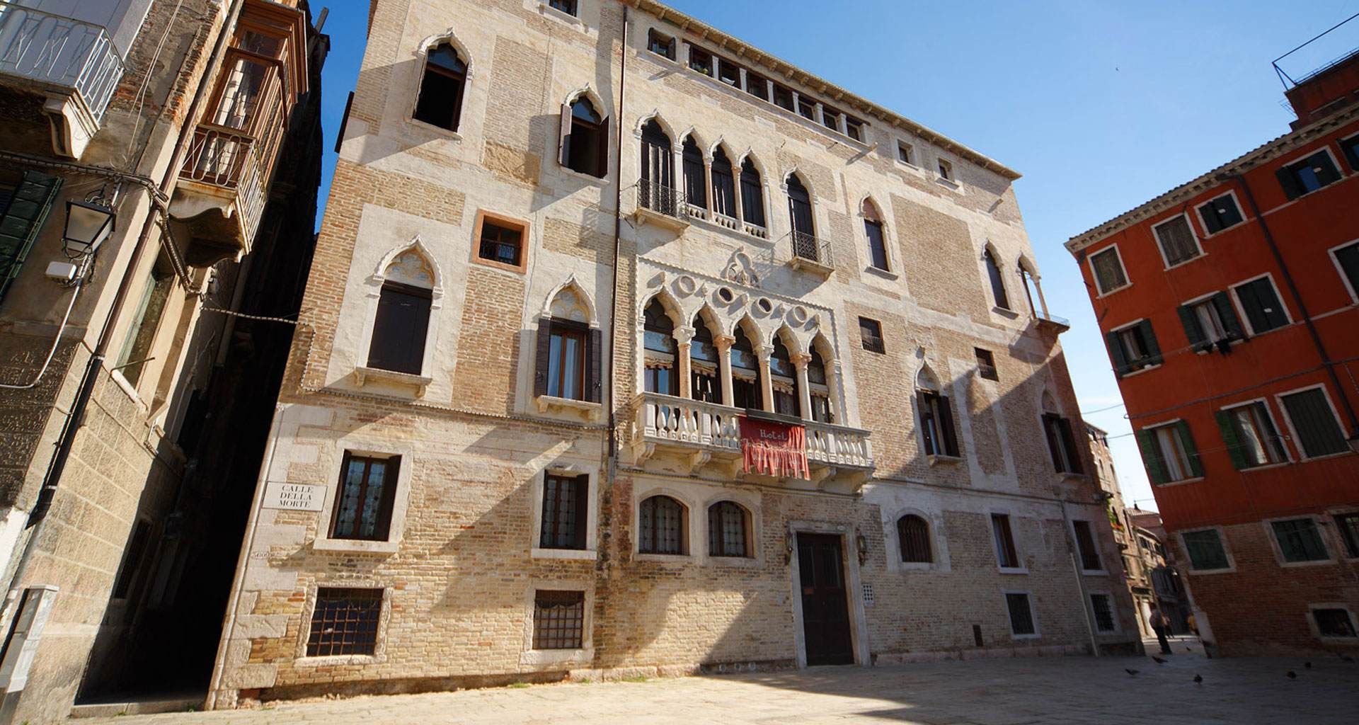 Cortile in pietra con un edificio storico color beige, finestre ad arco e balconi sotto un cielo azzurro.