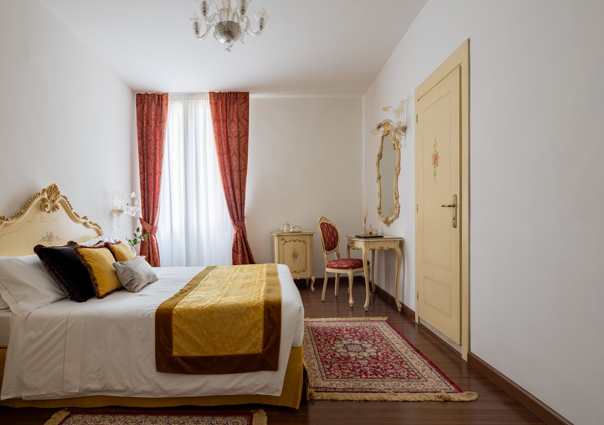 Elegante camera da letto con letto bianco, dettagli dorati, tende rosse, toletta, specchio e tappeto decorato.