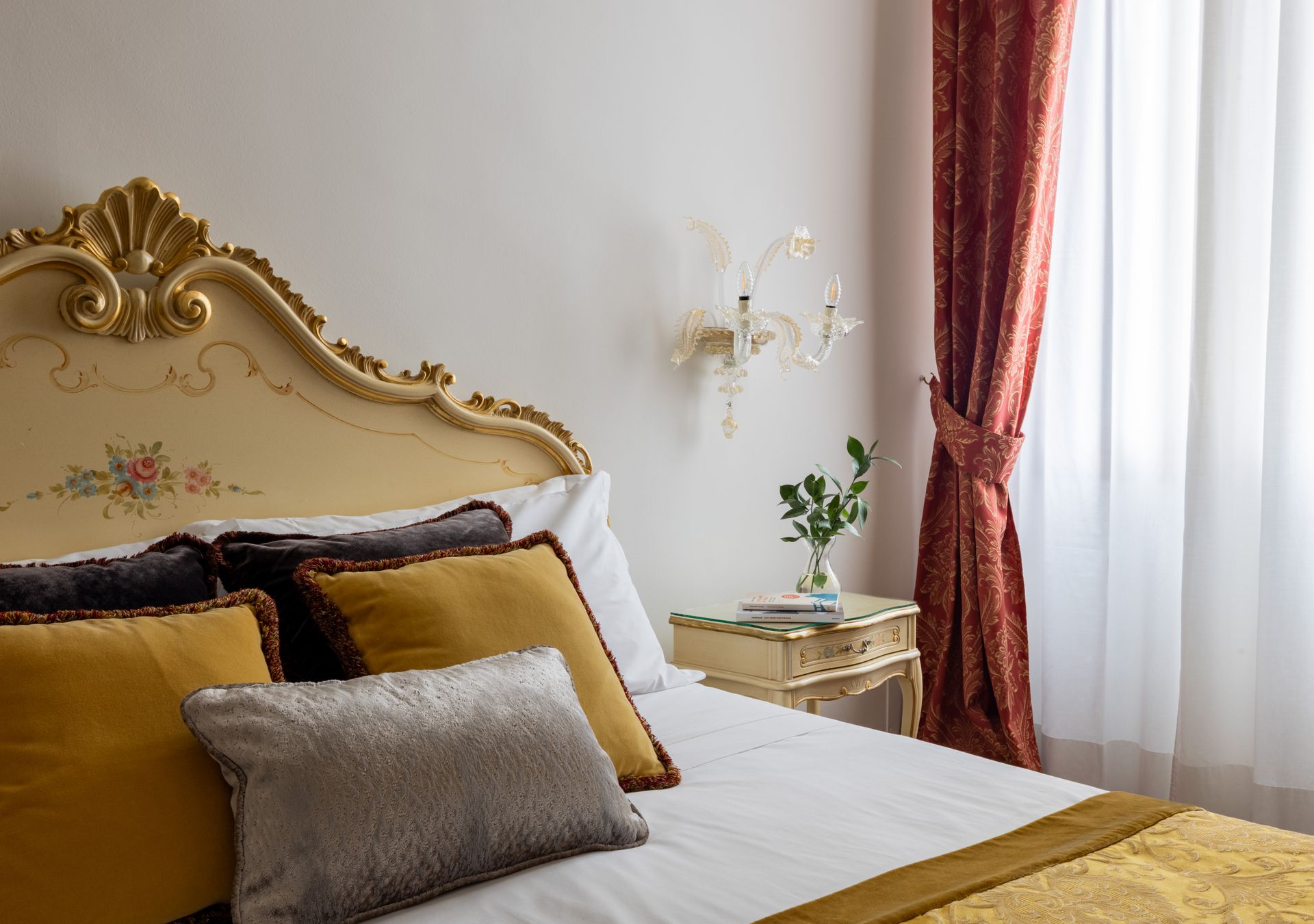 Elegante camera da letto con testiera dorata decorata, lenzuola bianche, cuscini color senape e tende rosse
