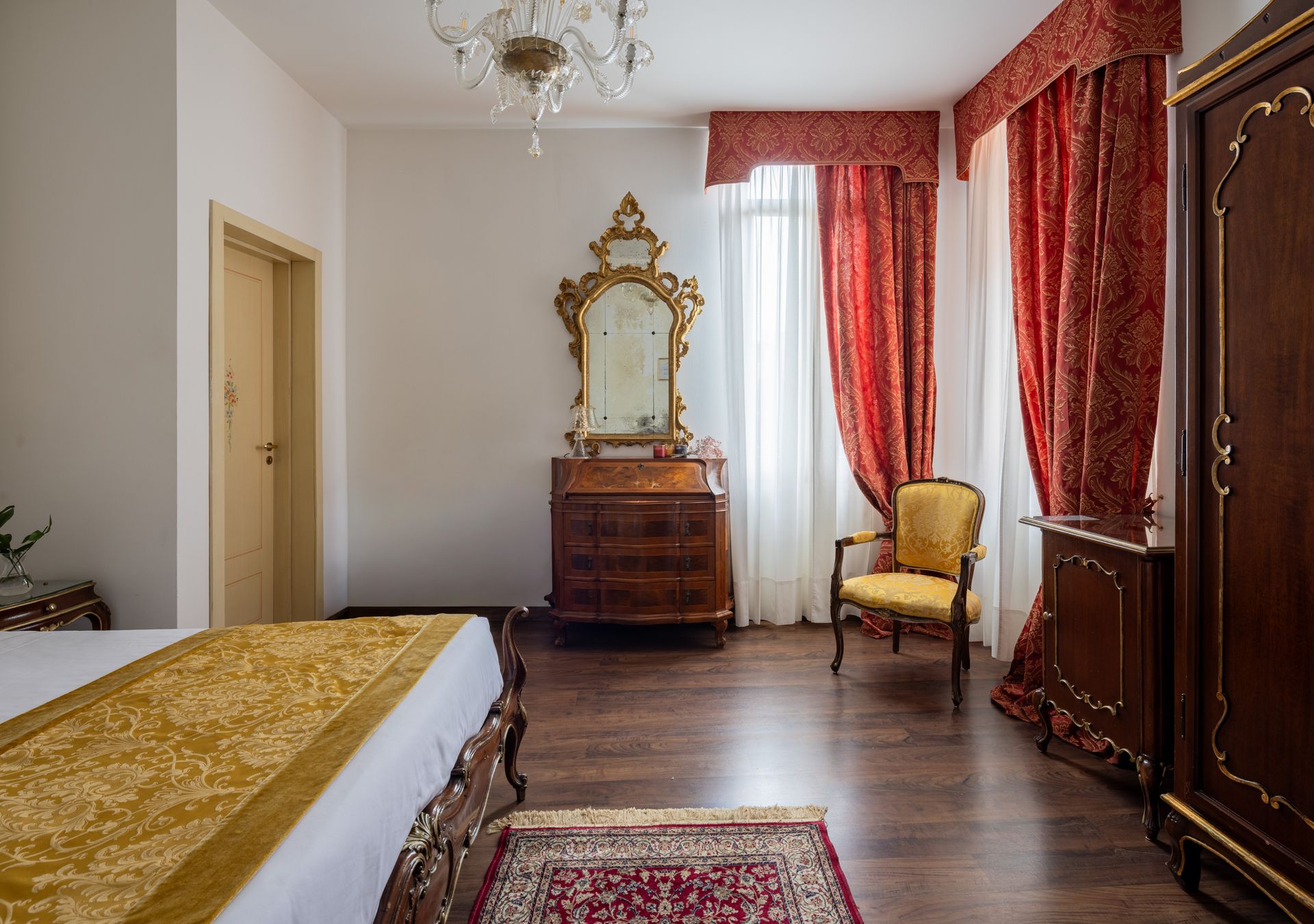 Elegante camera da letto con comò e specchio decorati, tende rosse, armadio in legno e poltrona imbottita.