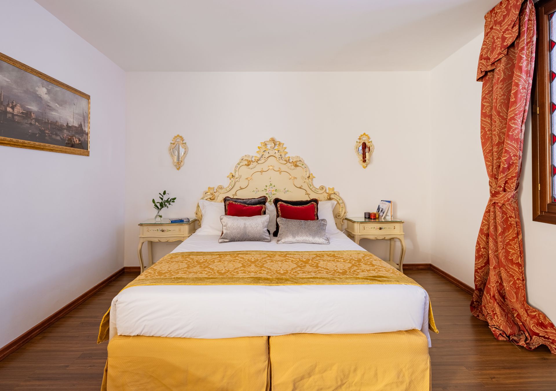 Elegante camera da letto con testiera dorata decorata, letto bianco, cuscini rossi e tende arancioni.