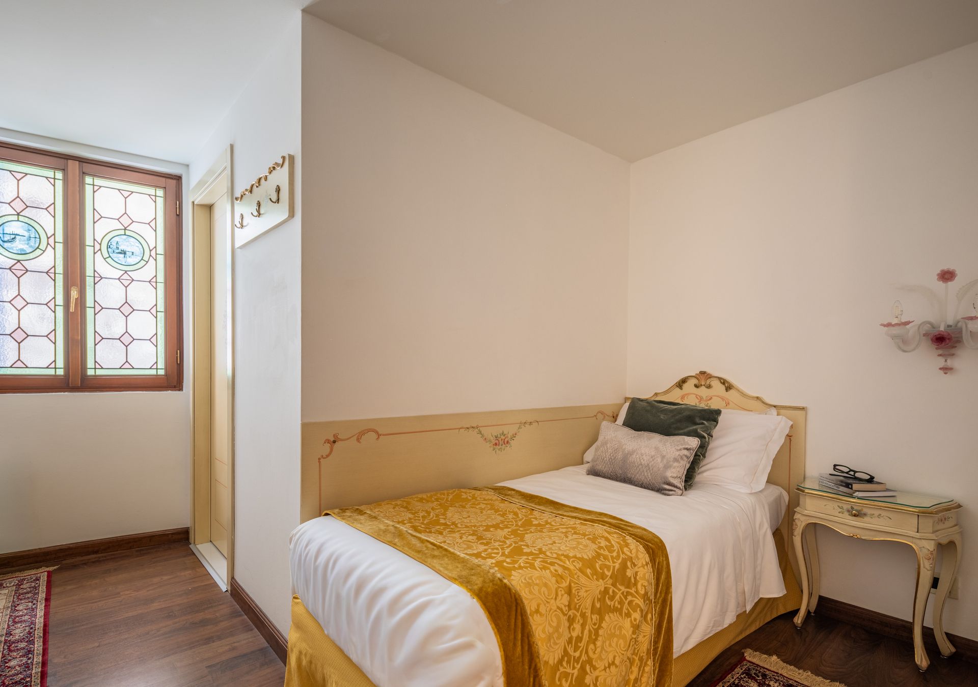 Piccola camera da letto con letto singolo, pareti bianche, lenzuola gialle, comodino e finestra con vetro decorato.