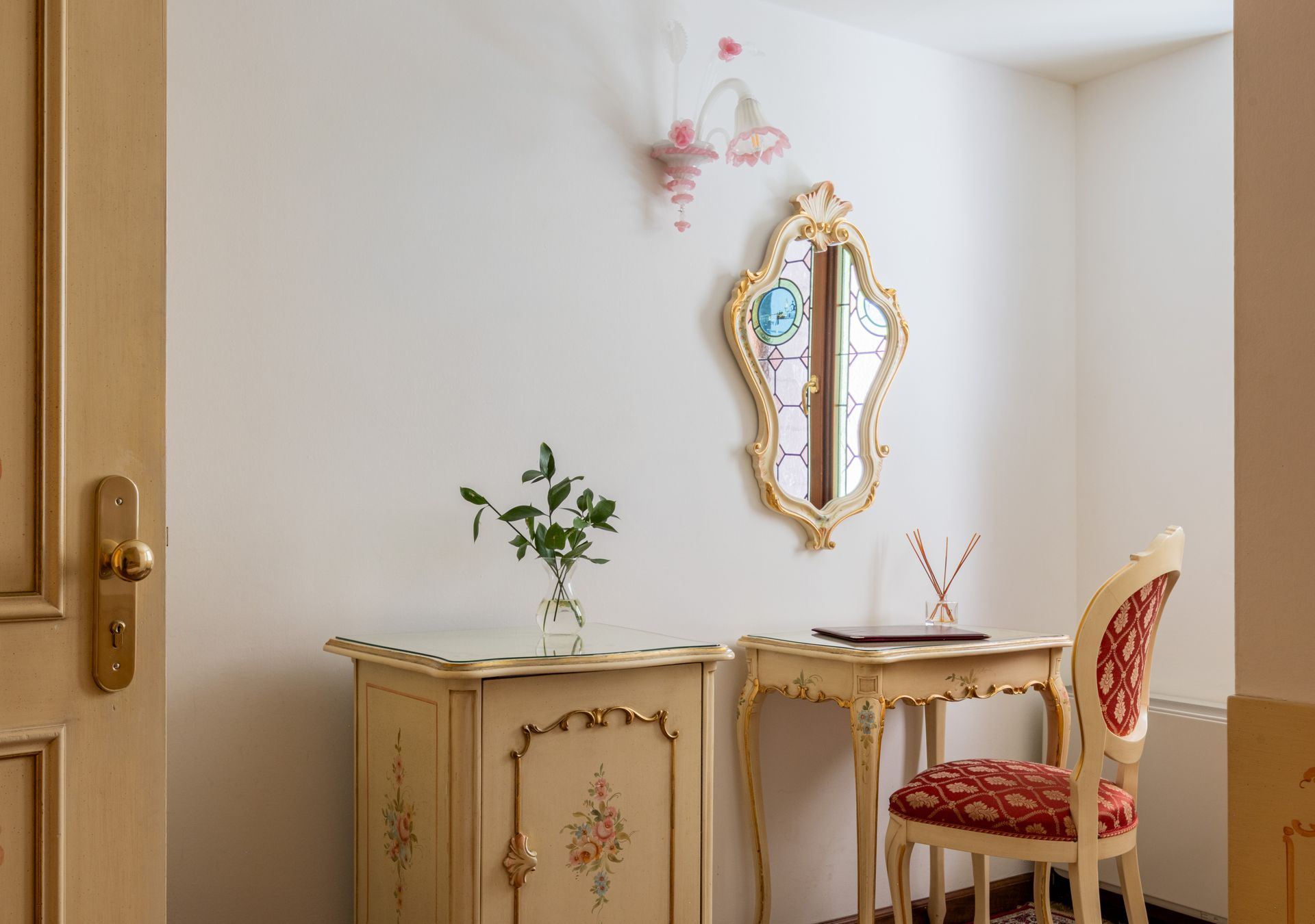 Elegante angolo con specchio decorato, piccola scrivania, sedia floreale e mobiletto in una stanza luminosa.