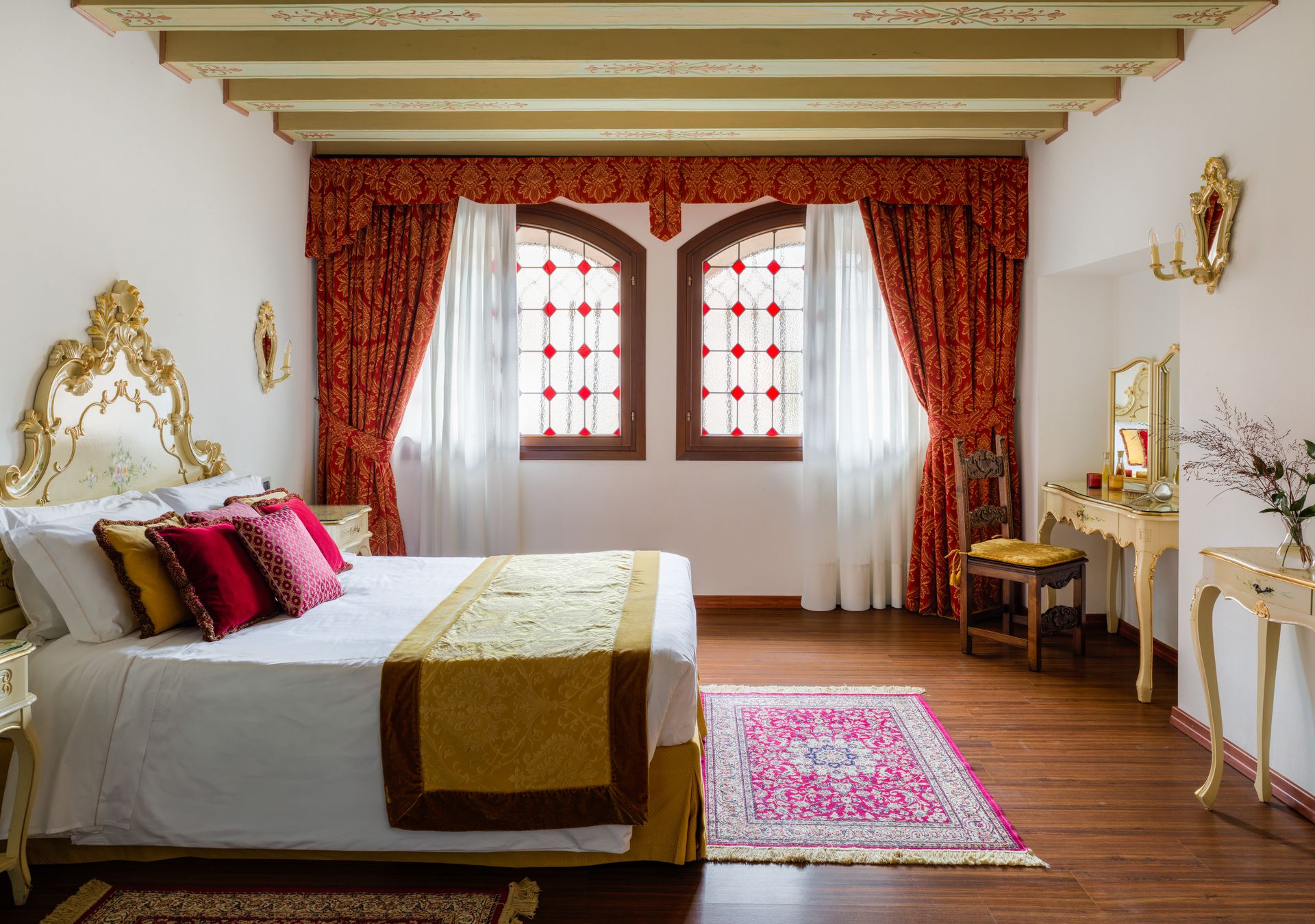 Elegante camera da letto con tende rosse, letto, pavimento in legno e tappeto rosa accanto a finestre ad arco.