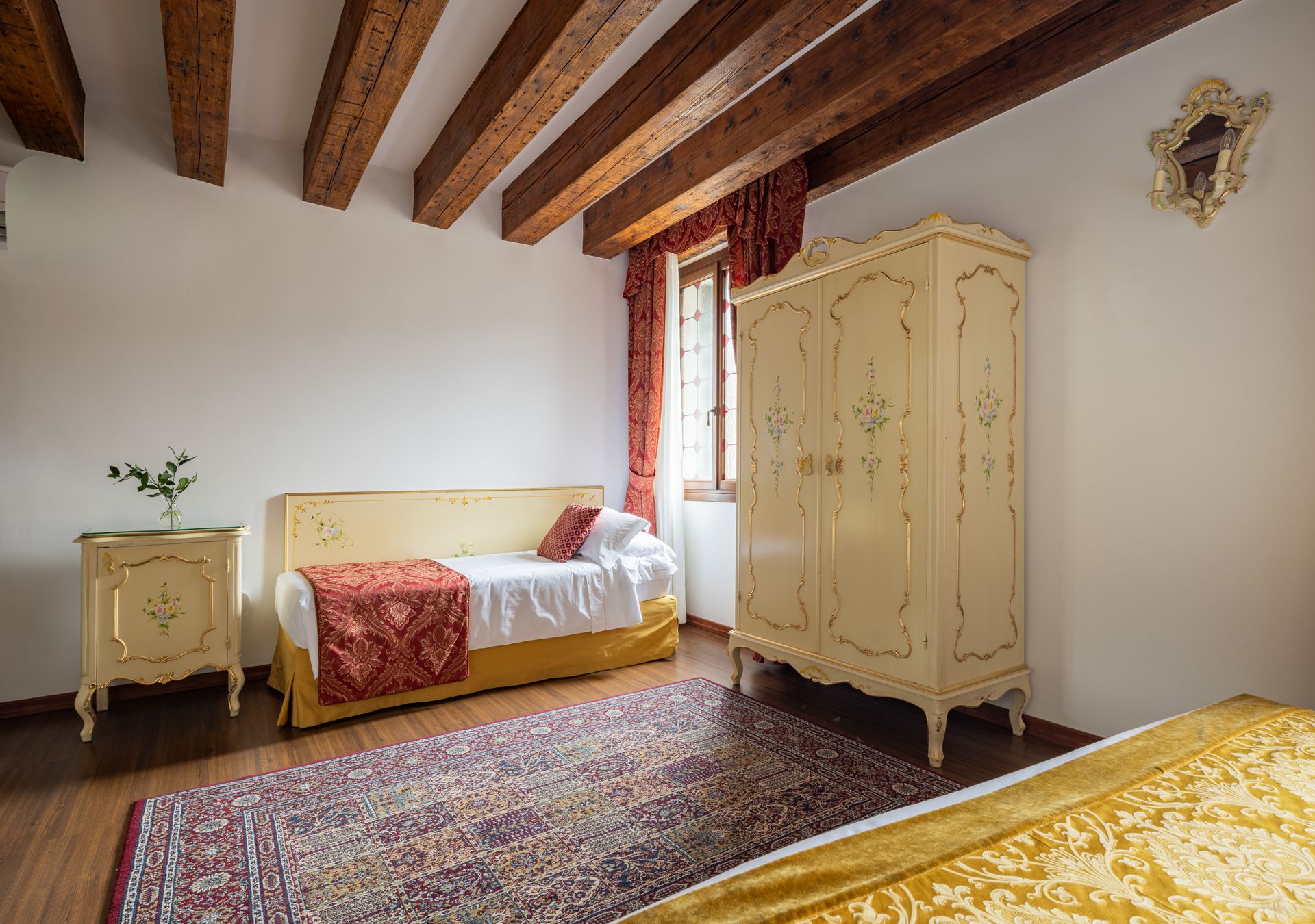 Camera da letto rustica con soffitto con travi a vista, armadi color crema decorati e letto con tessuti a fantasia.