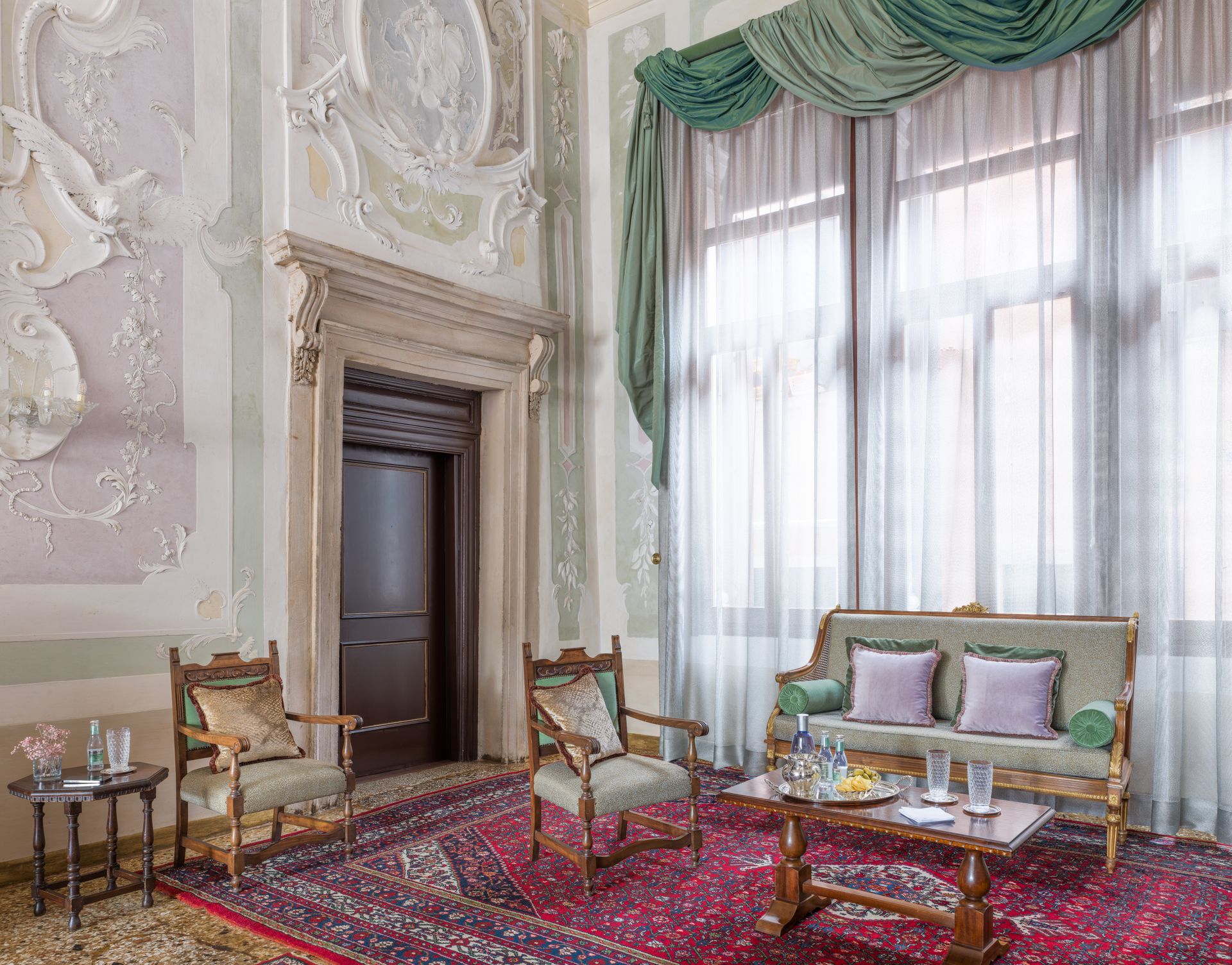 Elegante e sfarzoso salotto con alte finestre, sedie intagliate, divano e tappeto a motivi rossi.
