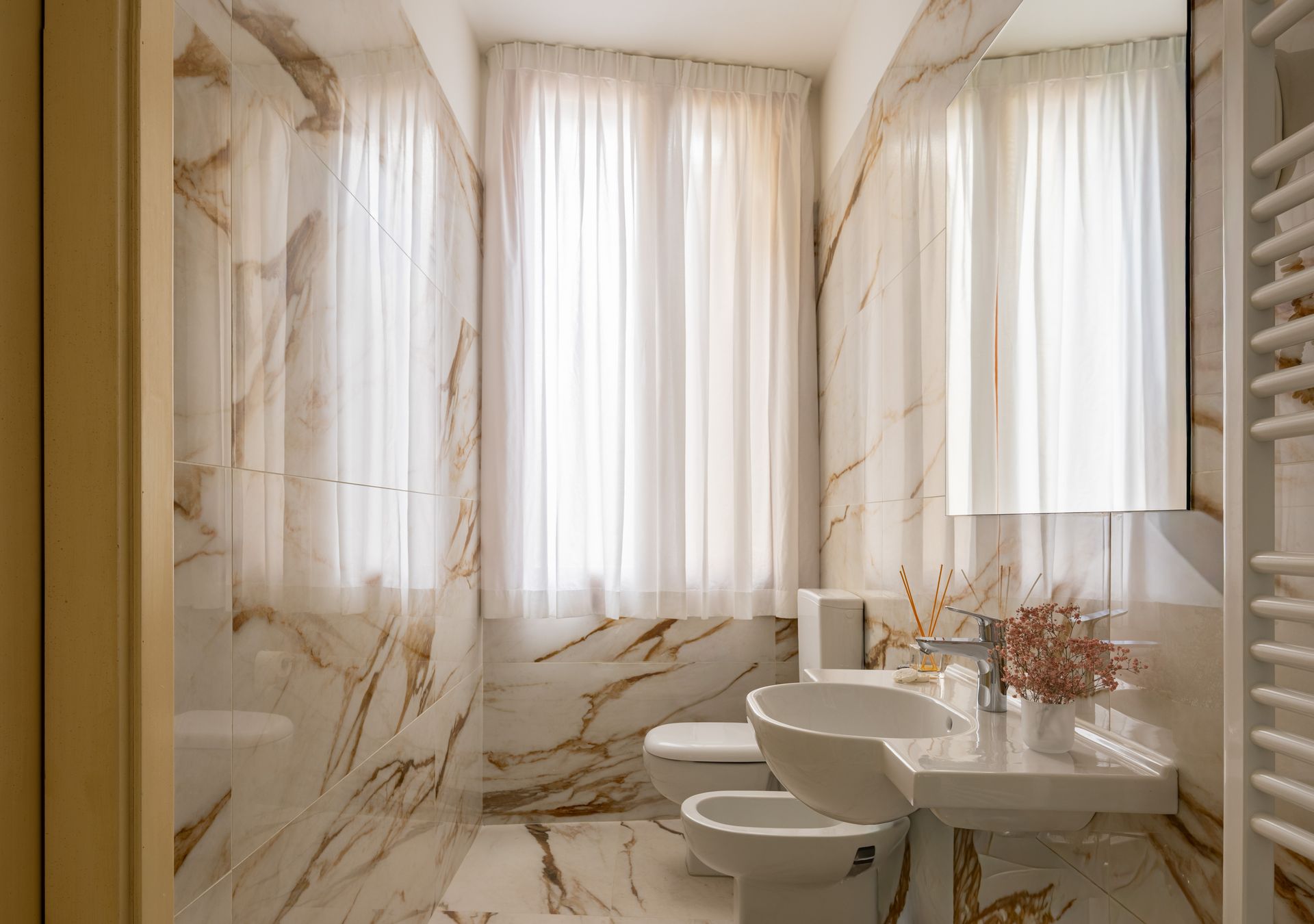 Elegante bagno in marmo con vasca, lavabo, WC e tende bianche trasparenti accanto a una finestra luminosa.
