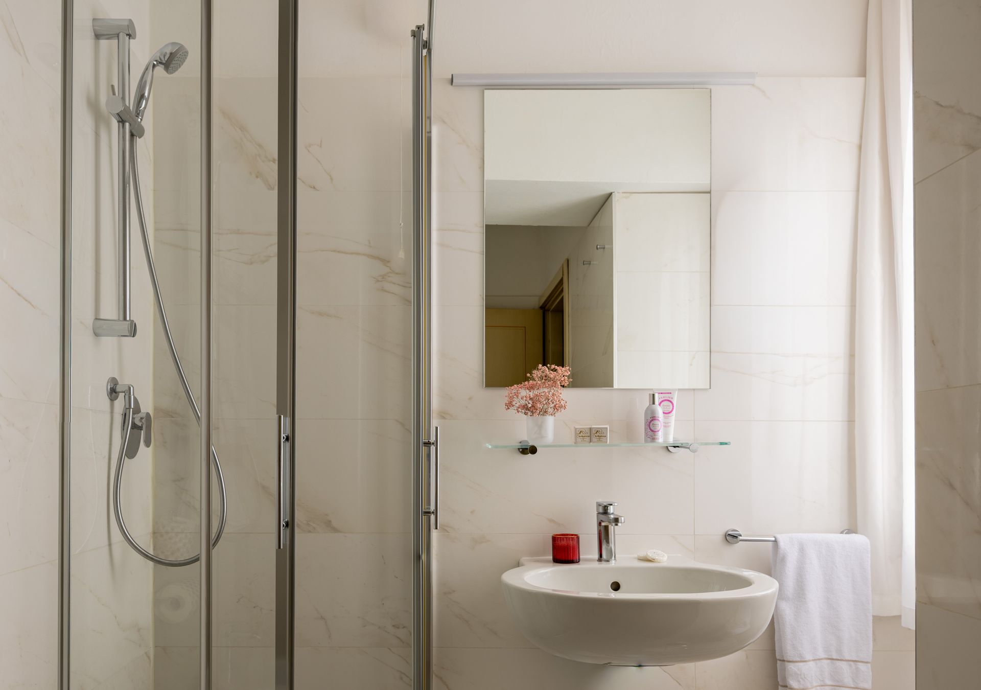 Bagno luminoso con doccia in vetro, specchio a parete, lavabo bianco e portasciugamani.