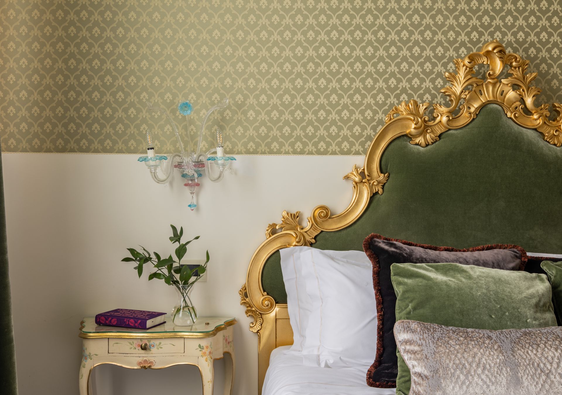 Elegante camera da letto con testiera dorata decorata, carta da parati a motivi e un piccolo comodino con fiori.
