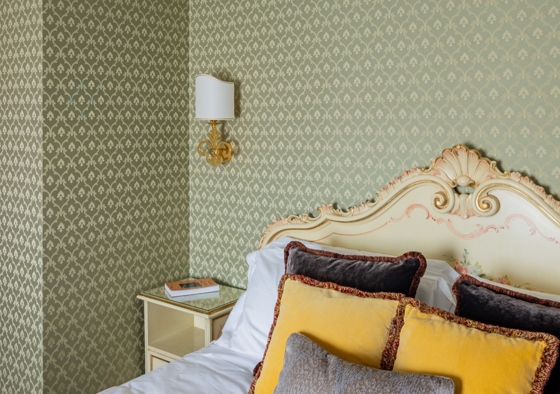 Elegante angolo camera da letto con carta da parati verde a motivi, testiera color crema e cuscini gialli.