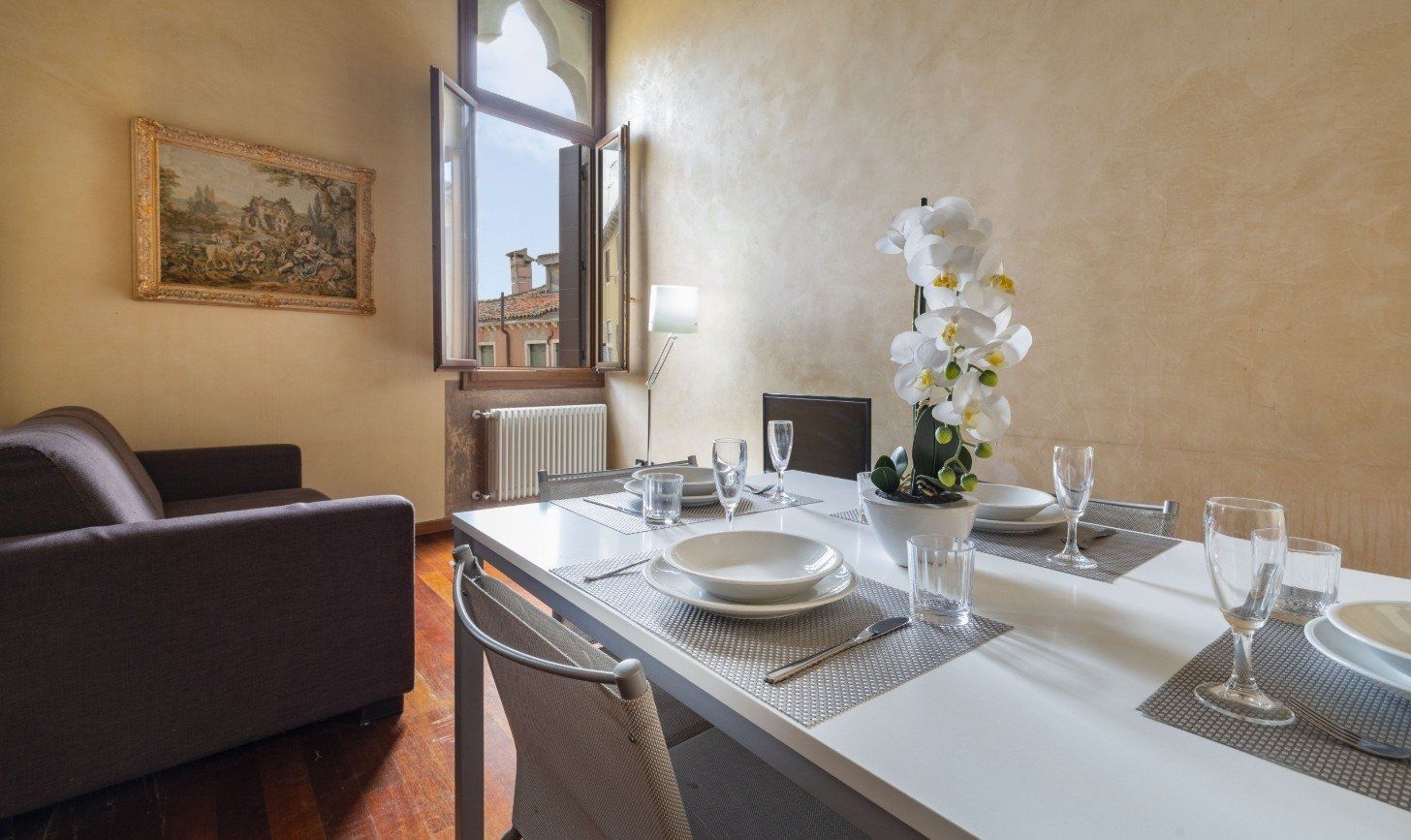 Salle à manger élégante avec table dressée, canapé foncé, fenêtre ouverte et fleurs sur un buffet clair.