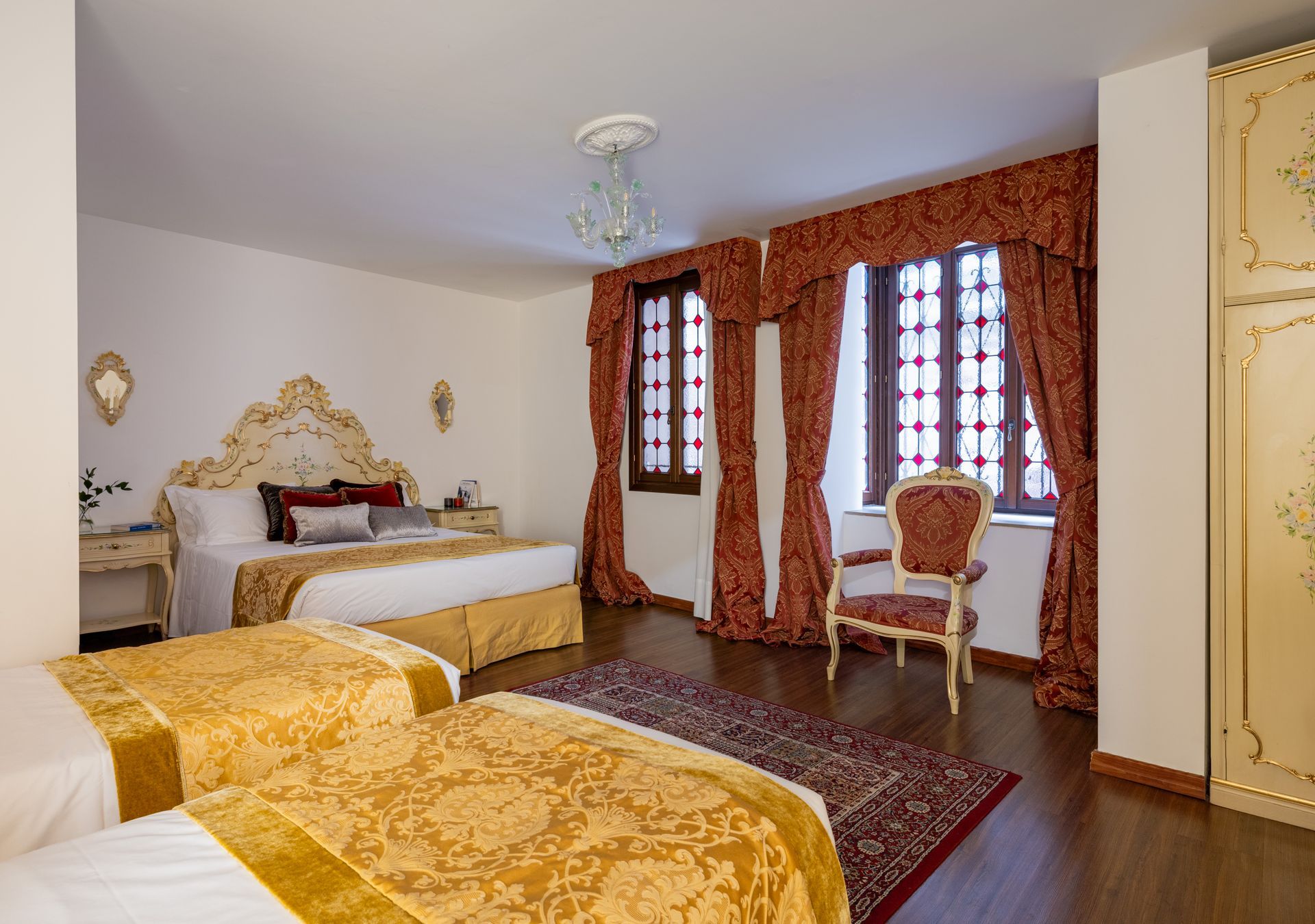Elegant bedroom with two beds, ornate gold accents, red curtains, and a chair by the window