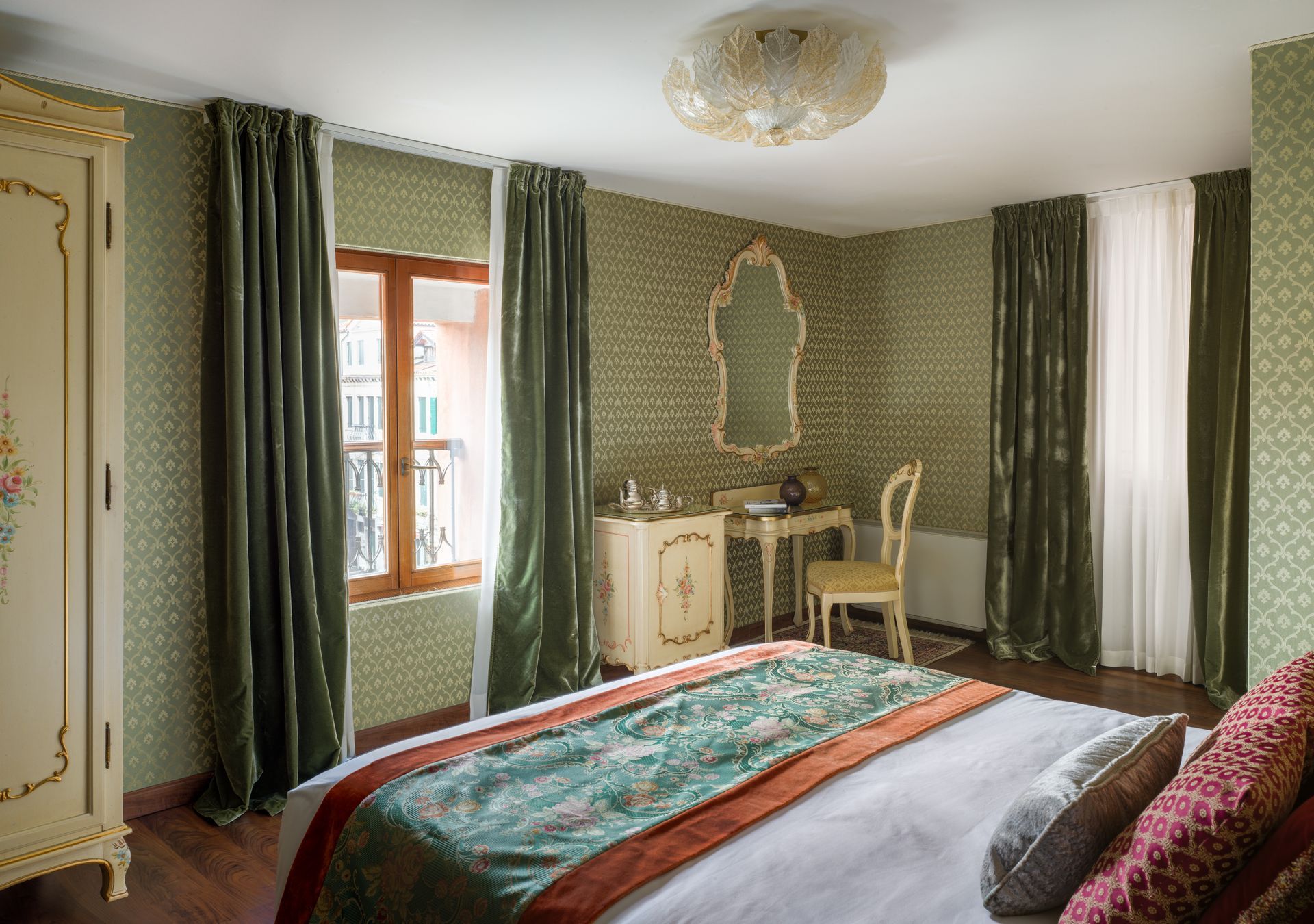 Elegant bedroom with green walls, ornate decor, desk, and a bed with red and teal bedding