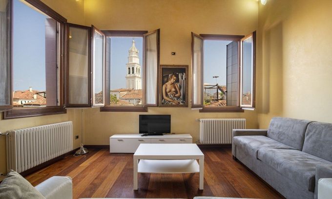 Bright living room with open windows, gray sofa, white coffee table, and city tower view.
