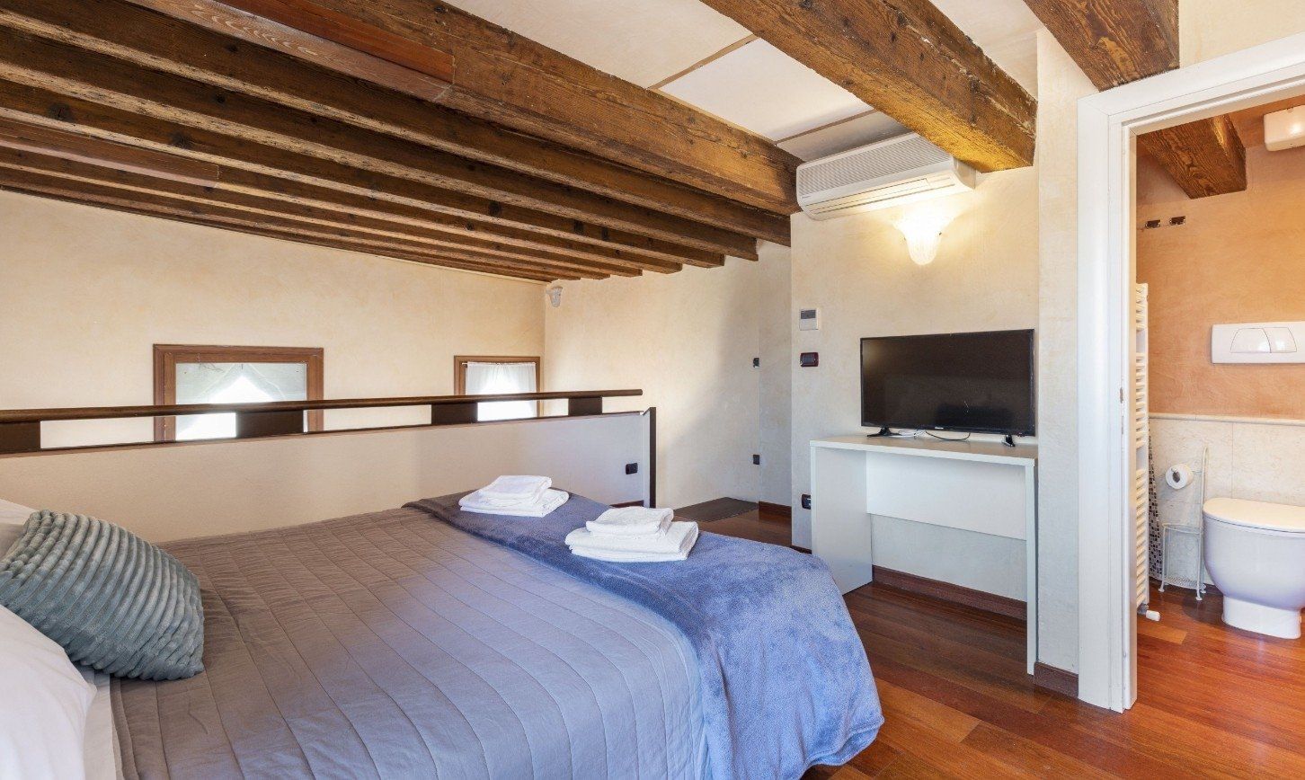 Chambre confortable avec poutres apparentes, couvre-lit gris et petit coin télé près de la porte