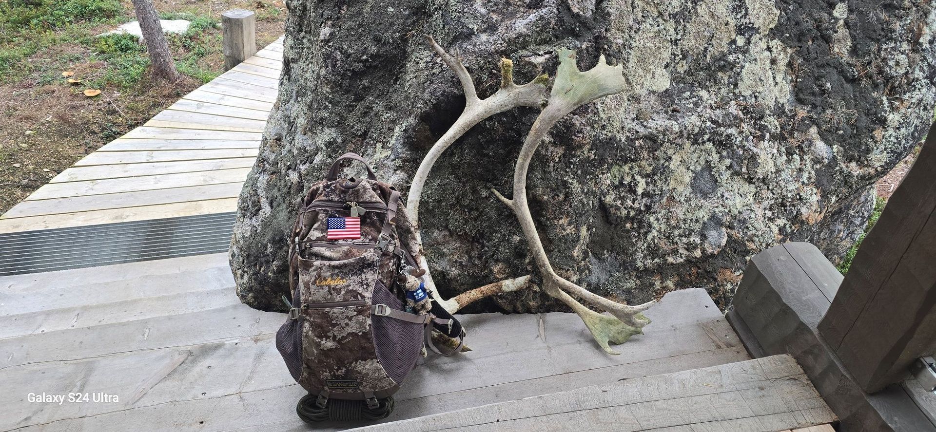 A camouflage backpack and a pair of antlers rest on stone steps near a rock.