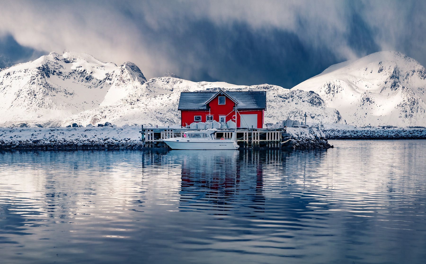 A red house is sitting in the middle of a lake surrounded by snow covered mountains.