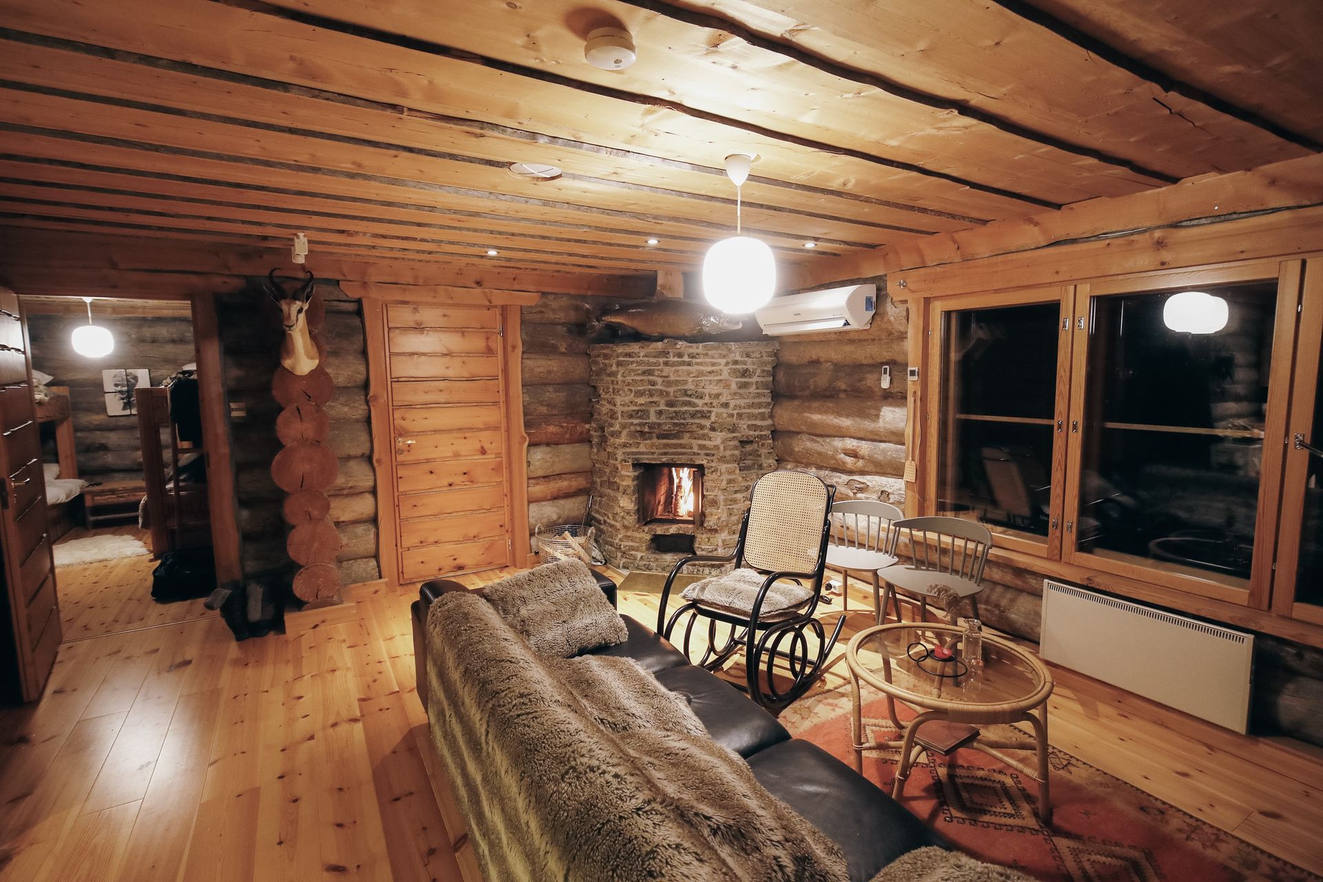 Warm cabin living room with wooden walls, stone fireplace, sofa, rocking chairs, and large windows