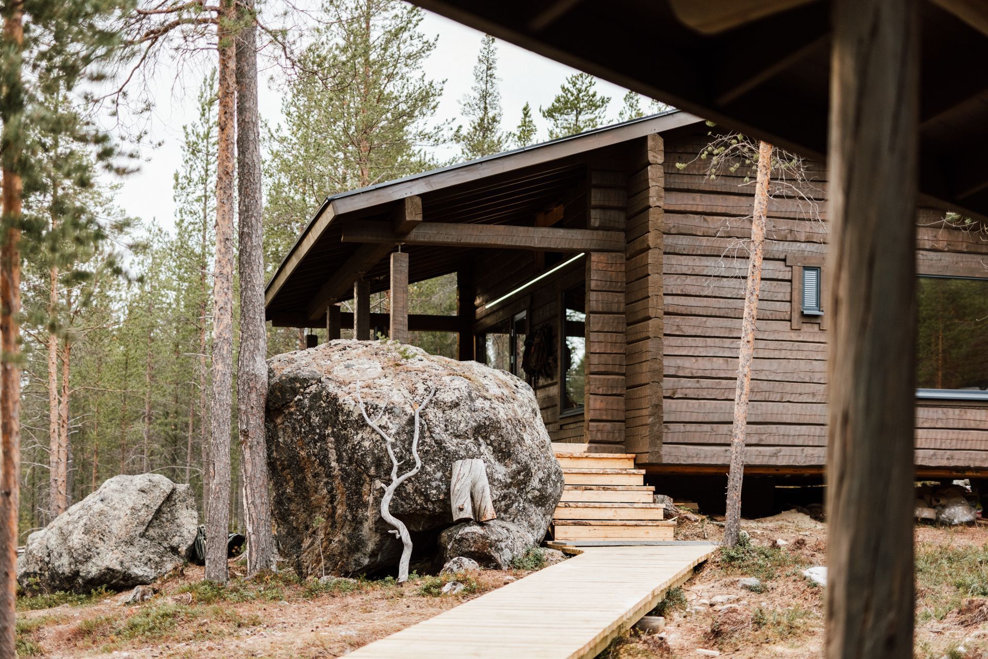 A wooden house in the middle of a forest with a large rock in front of it.