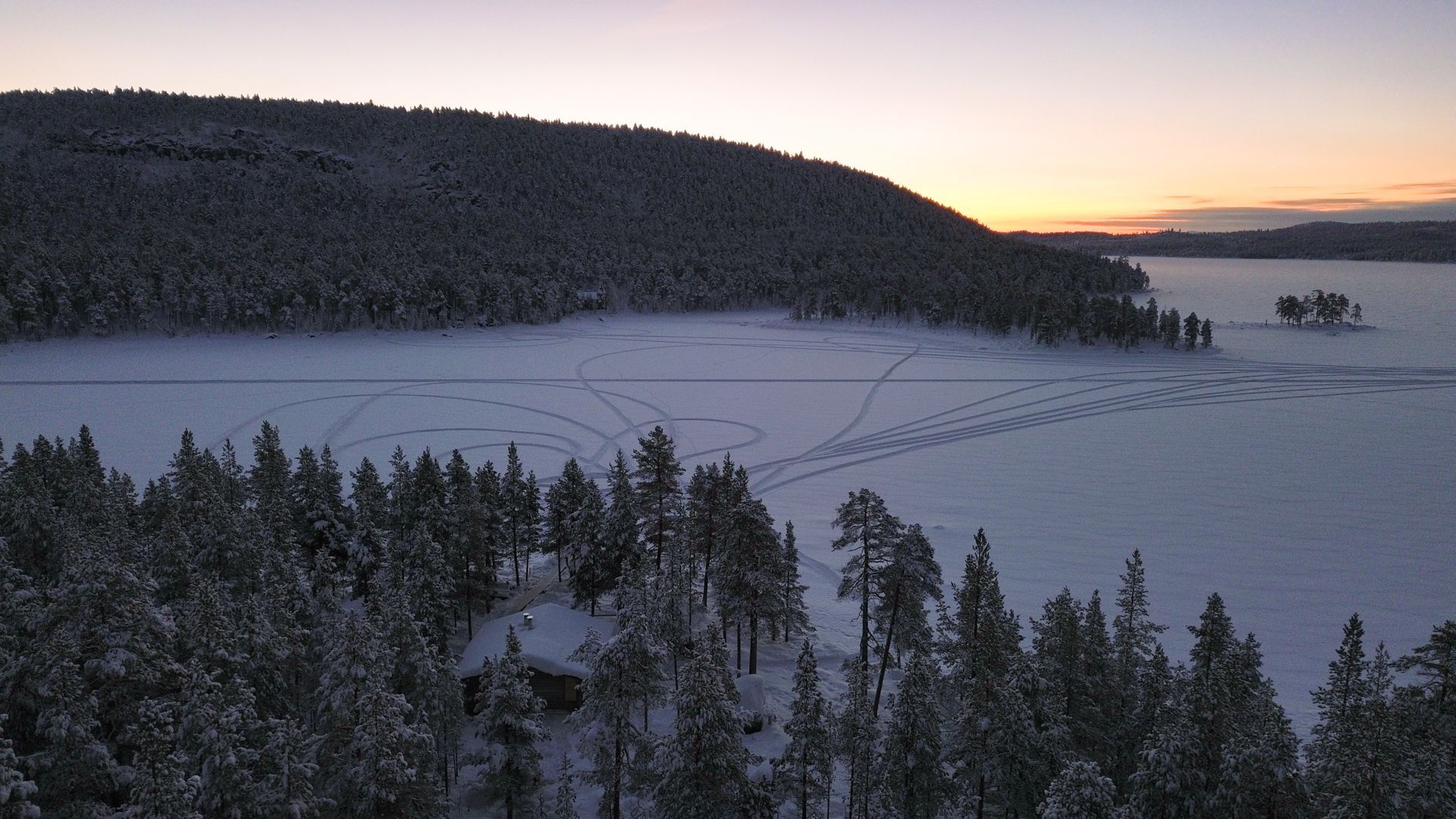 An aerial view of a frozen lake surrounded by snow covered trees at sunset.