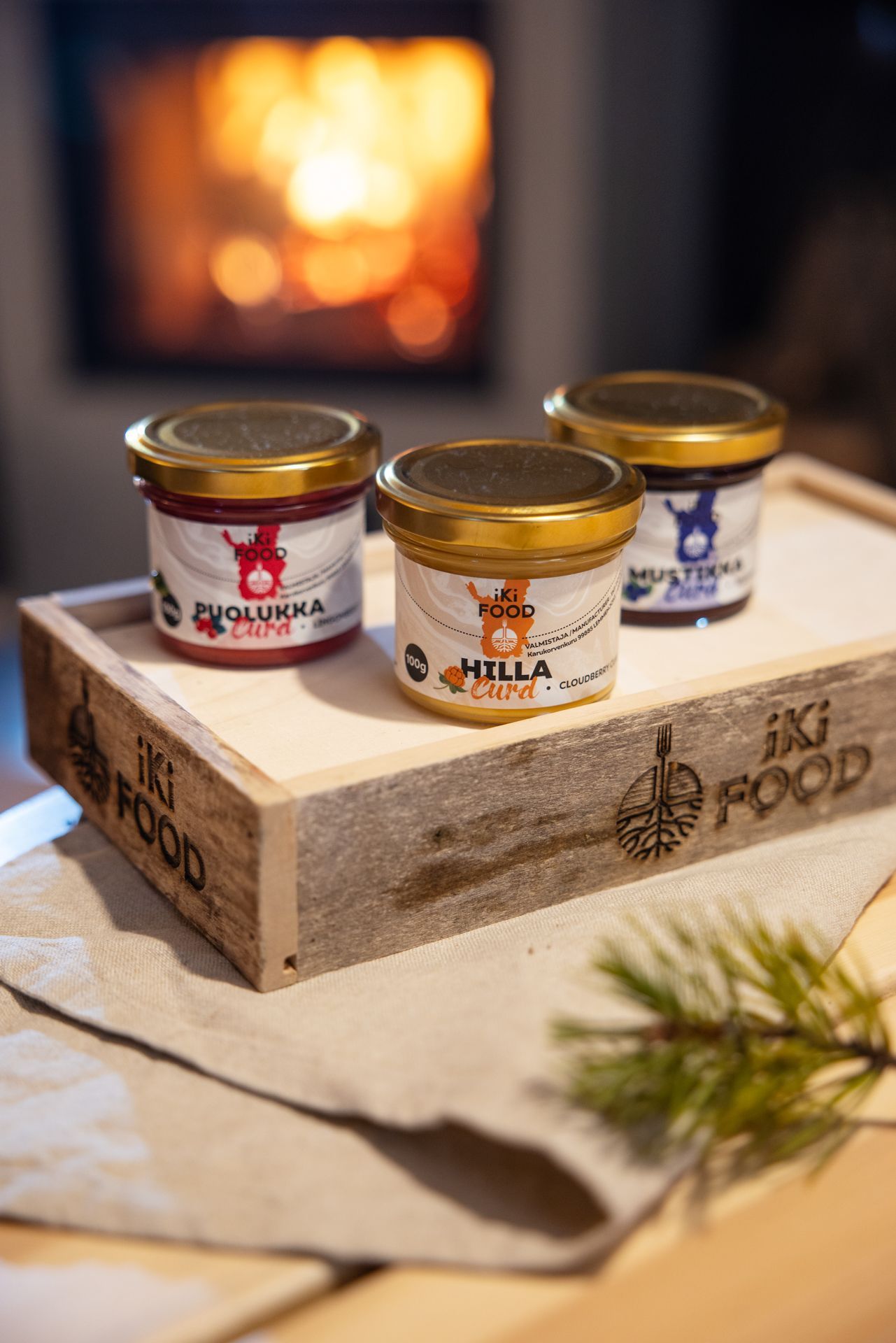 Three jars of food products on a wooden tray with a fireplace in the background.