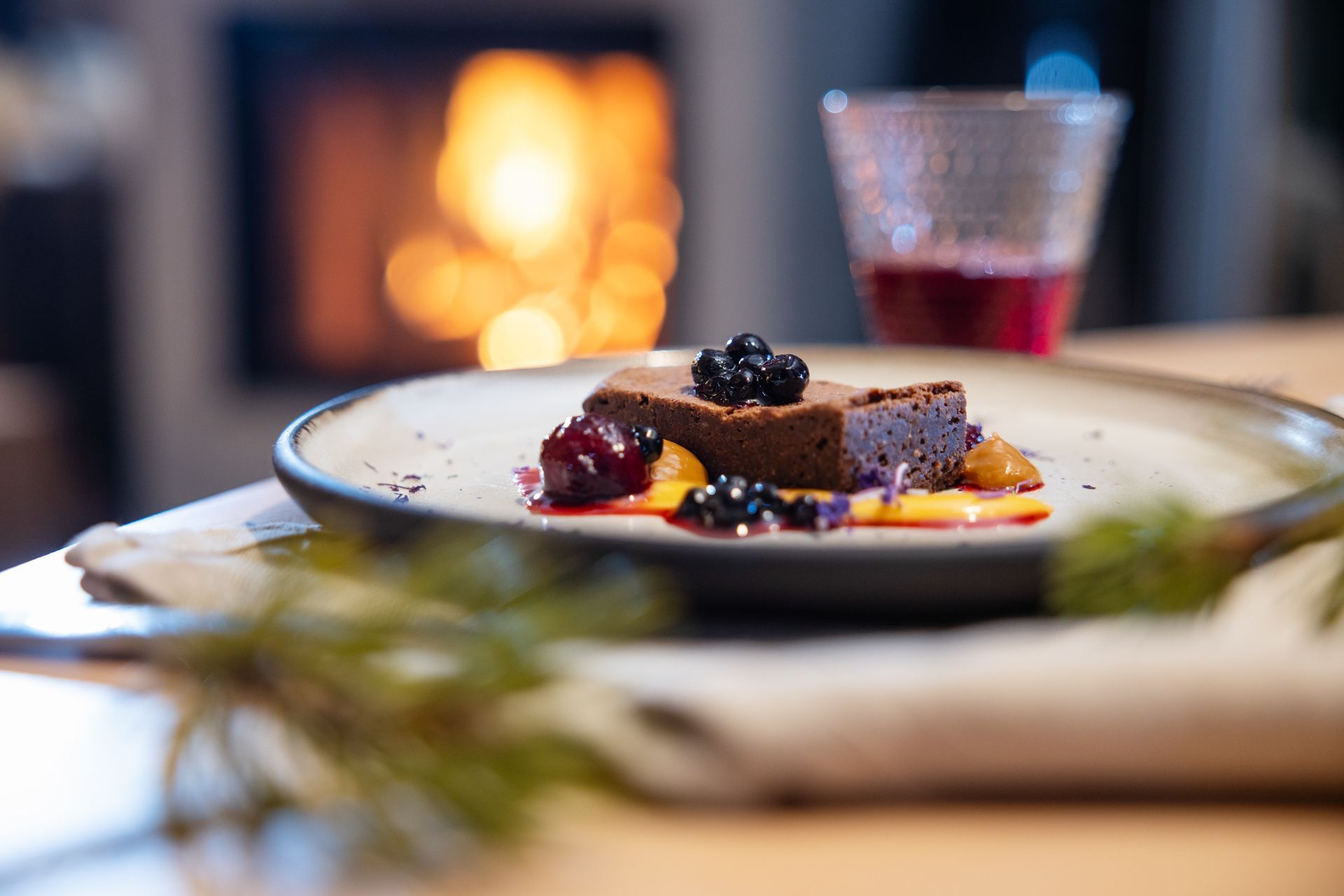 Brownie on plate, garnished with fruit, in front of a fireplace with drink.