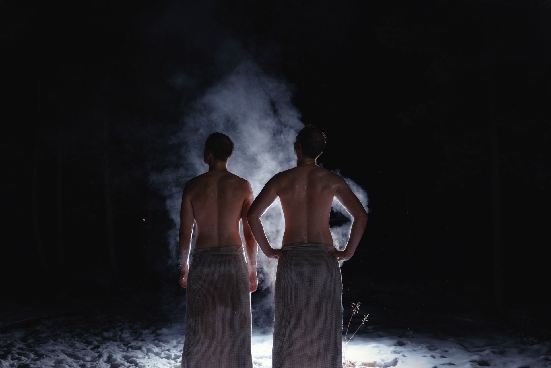 Two people wrapped in towels stand outdoors, back to camera, in front of a cloud of steam in the dark.