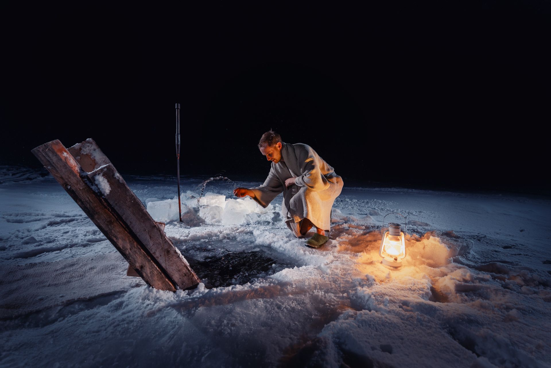 Man by ice hole at night, illuminated by lantern. He is using a bucket to scoop something.