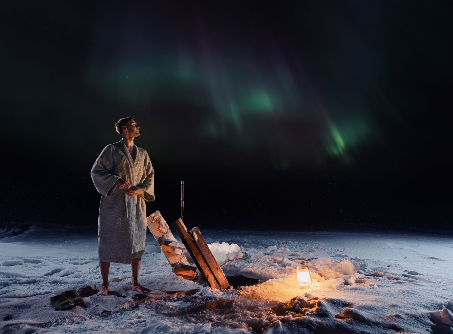 Person in robe looks up at aurora borealis over snowy ground, near fire and hole.