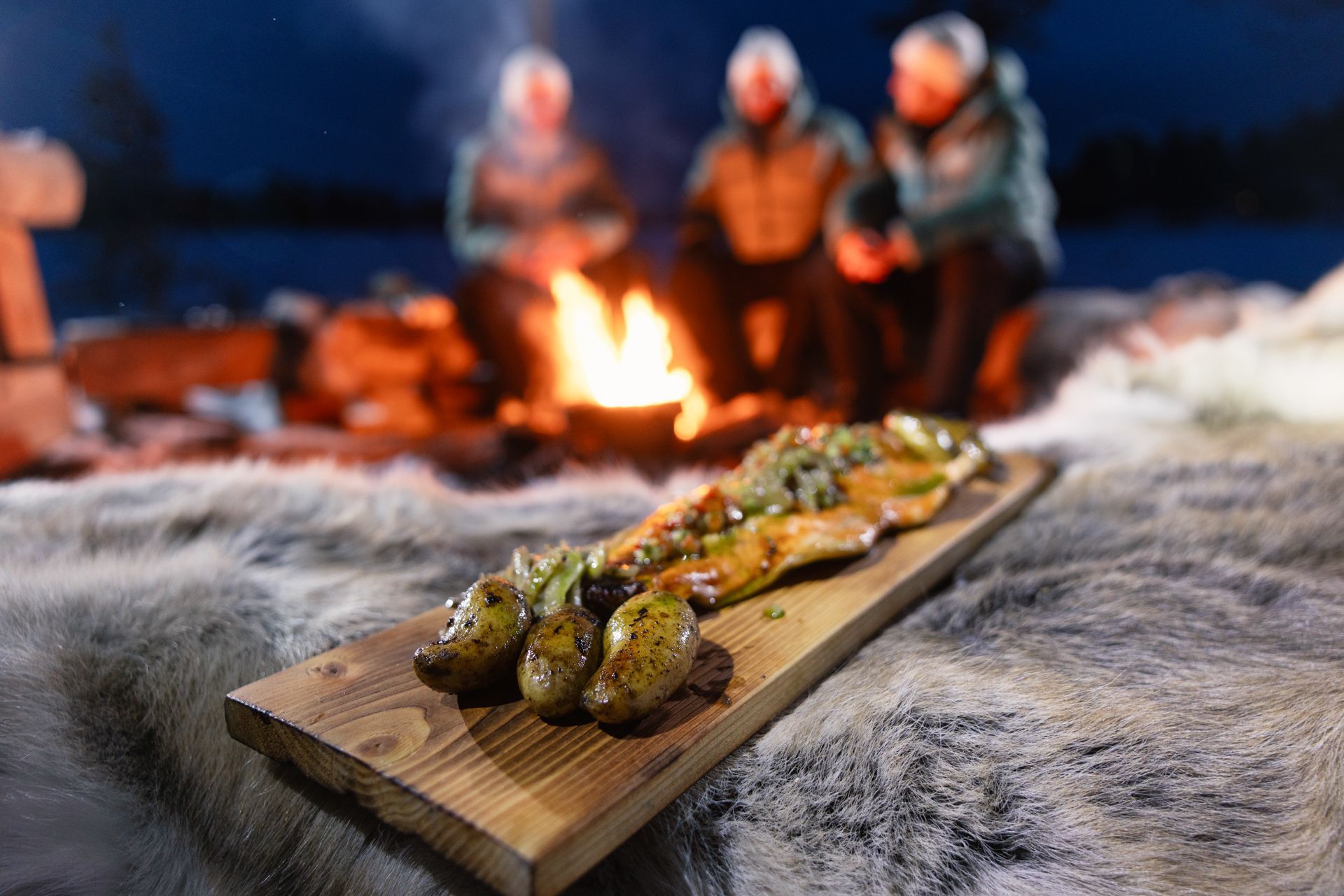 Grilled fish and potatoes on wooden board with people by a campfire in snow.