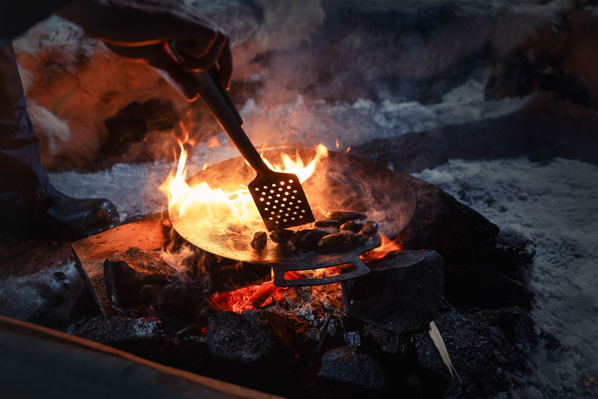Person cooking over an open fire, stirring food in a pan with a slotted spatula.