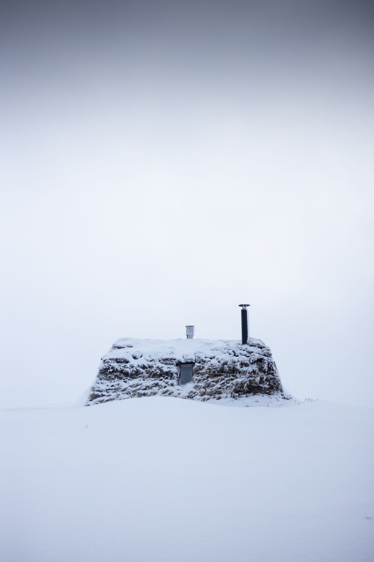 Snow-covered stone hut with chimney and door against a white, overcast sky.