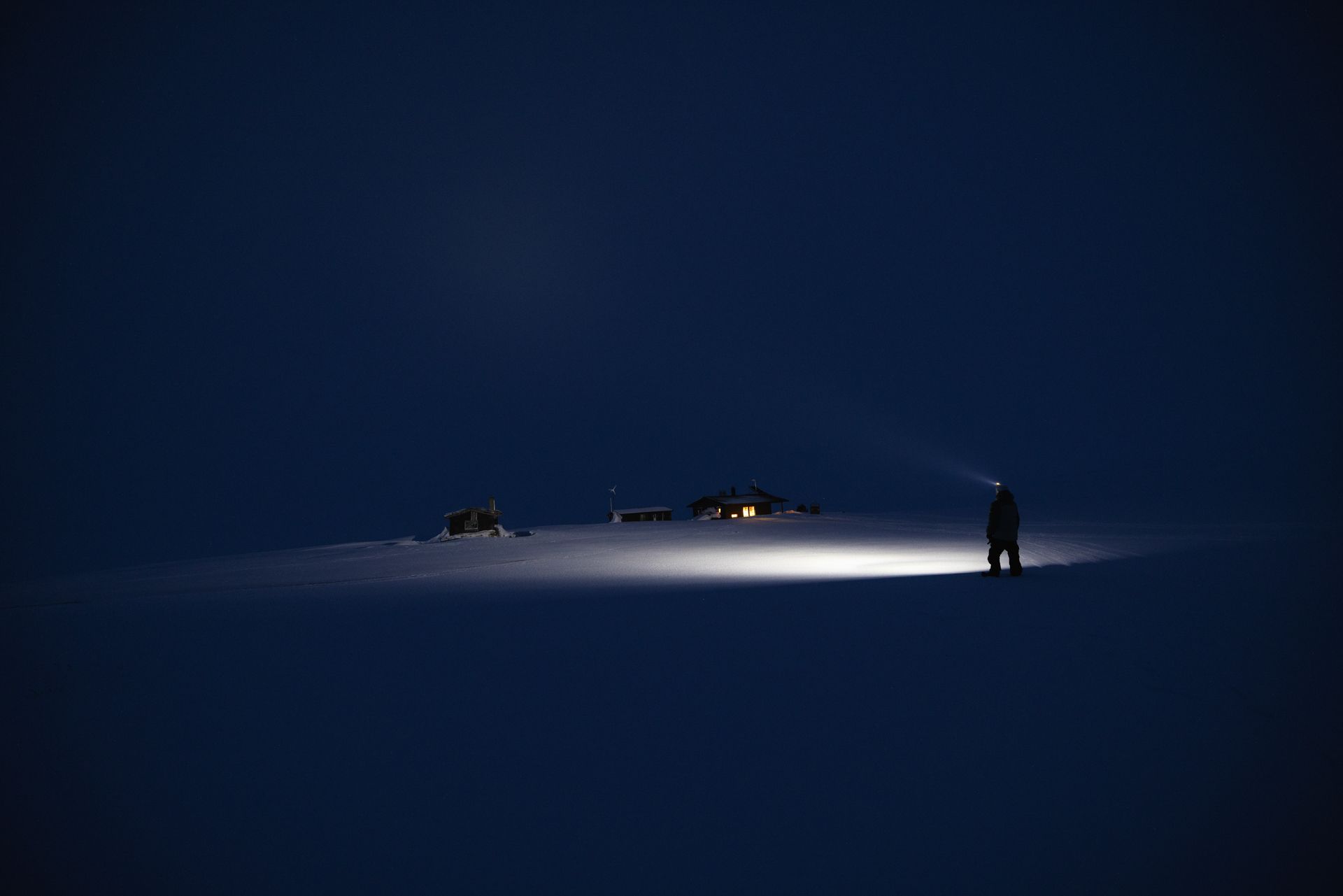 Person in the snow at night, wearing a headlamp. A cabin is dimly lit in the distance.