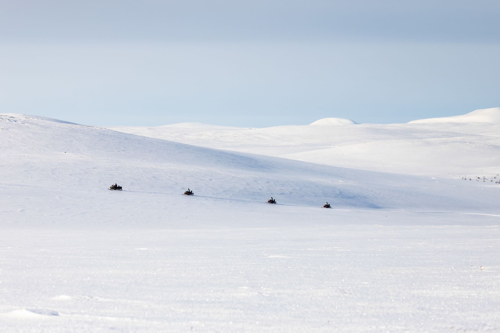 Snowmobiles driving across a vast, snow-covered landscape under a light blue sky.