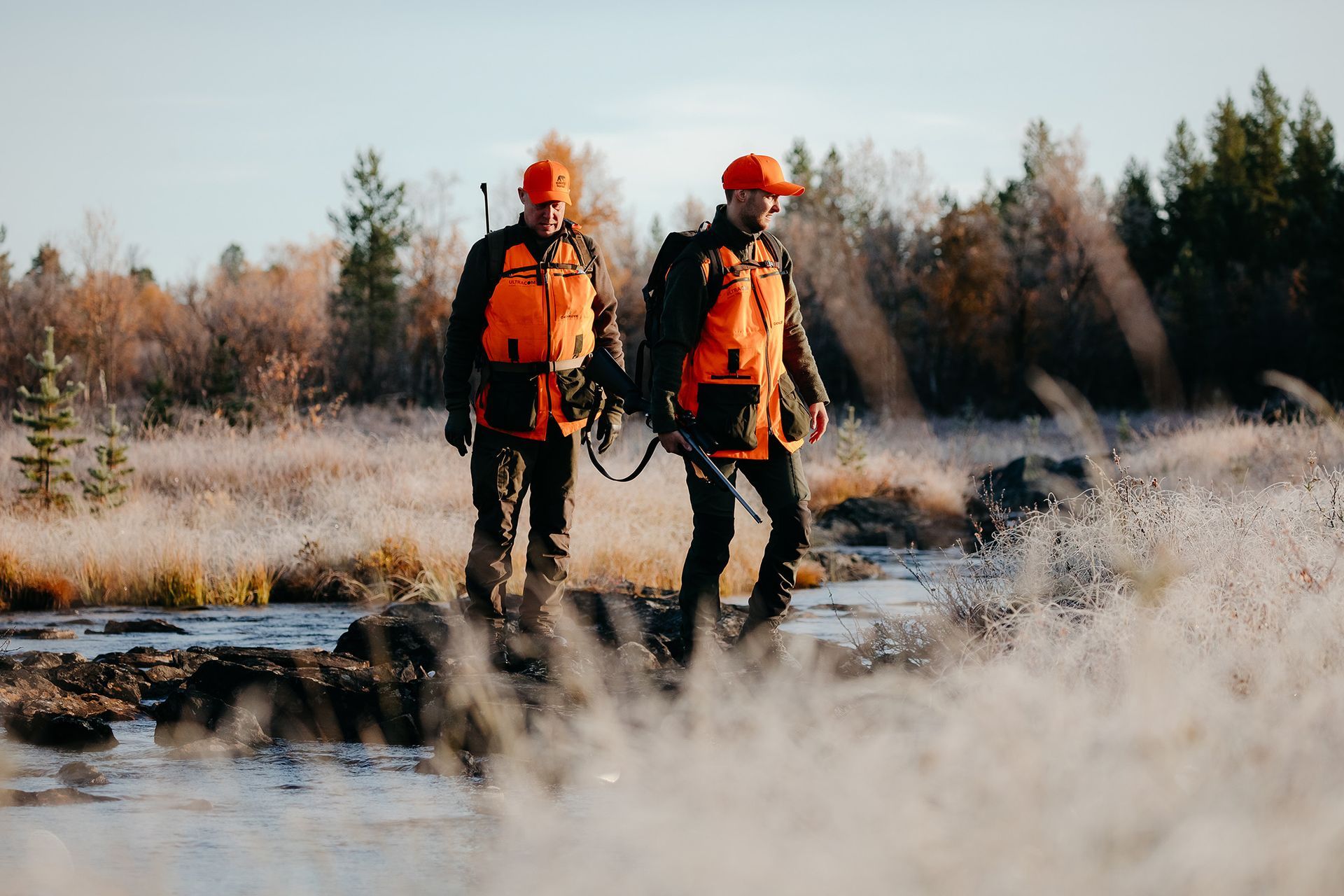 Two men in orange jackets are walking across a river.