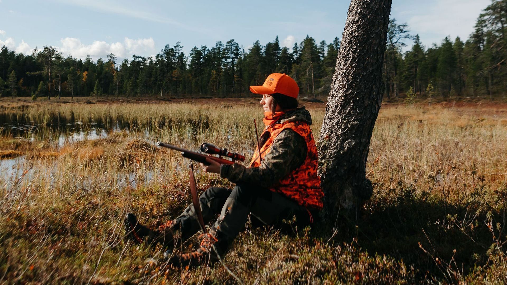 a woman sitting by a tree by a swamp holding a hunting riffle