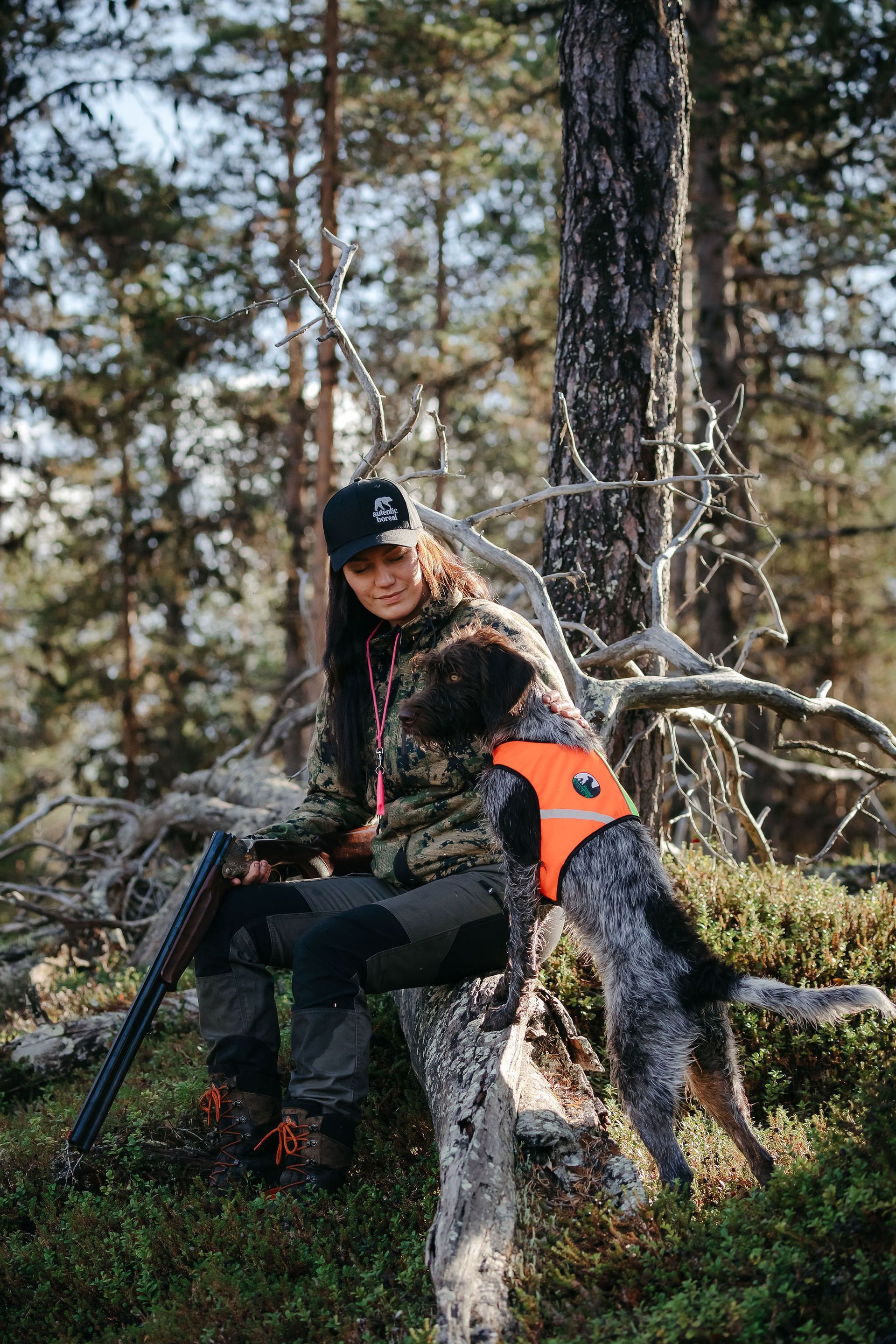 Woman in camouflage gear with a dog wearing an orange vest in a forest, holding a rifle.