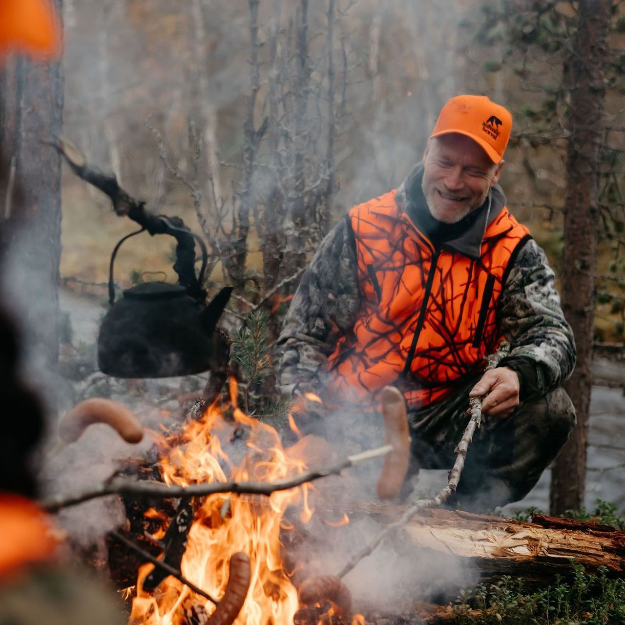 A man in an orange vest is kneeling around a campfire