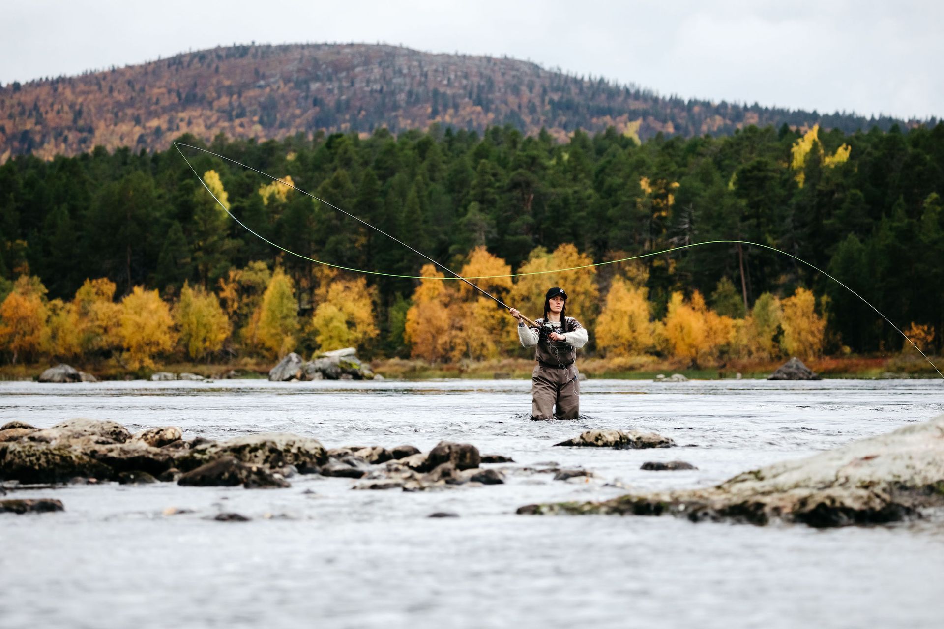 A man is fishing in a river with mountains in the background.