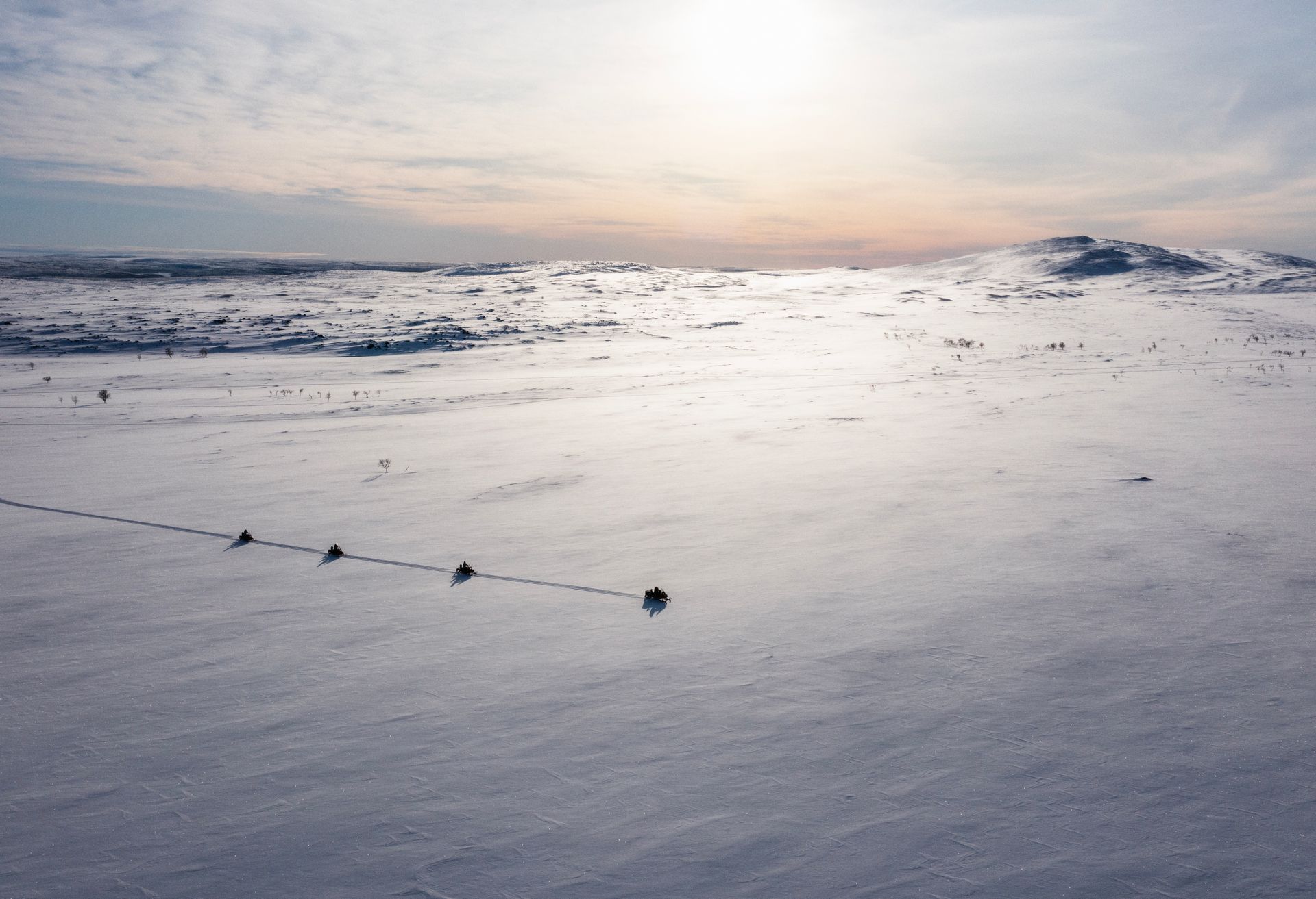 Snow-covered landscape with a line of small dark figures moving across it under a bright sky.