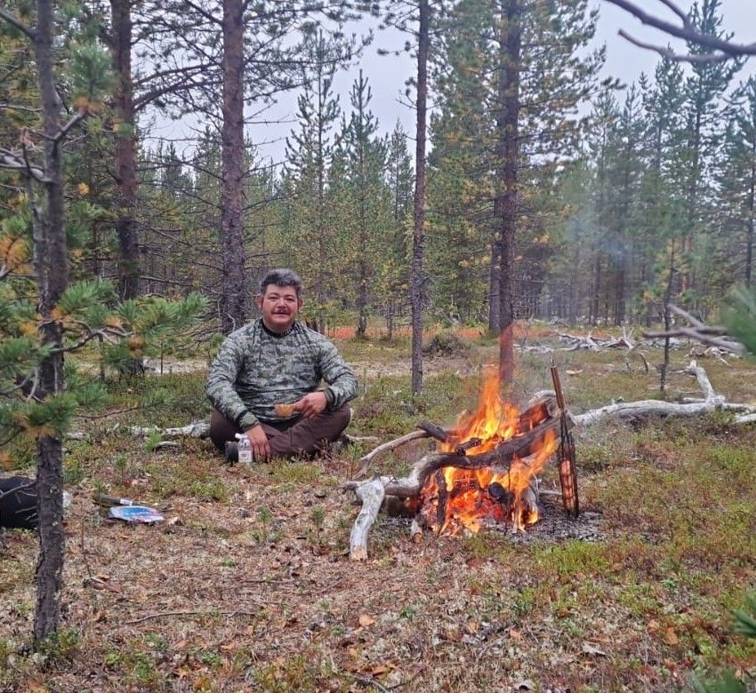 Man sitting near campfire in forest, holding item. Fall foliage, trees, and cloudy sky.