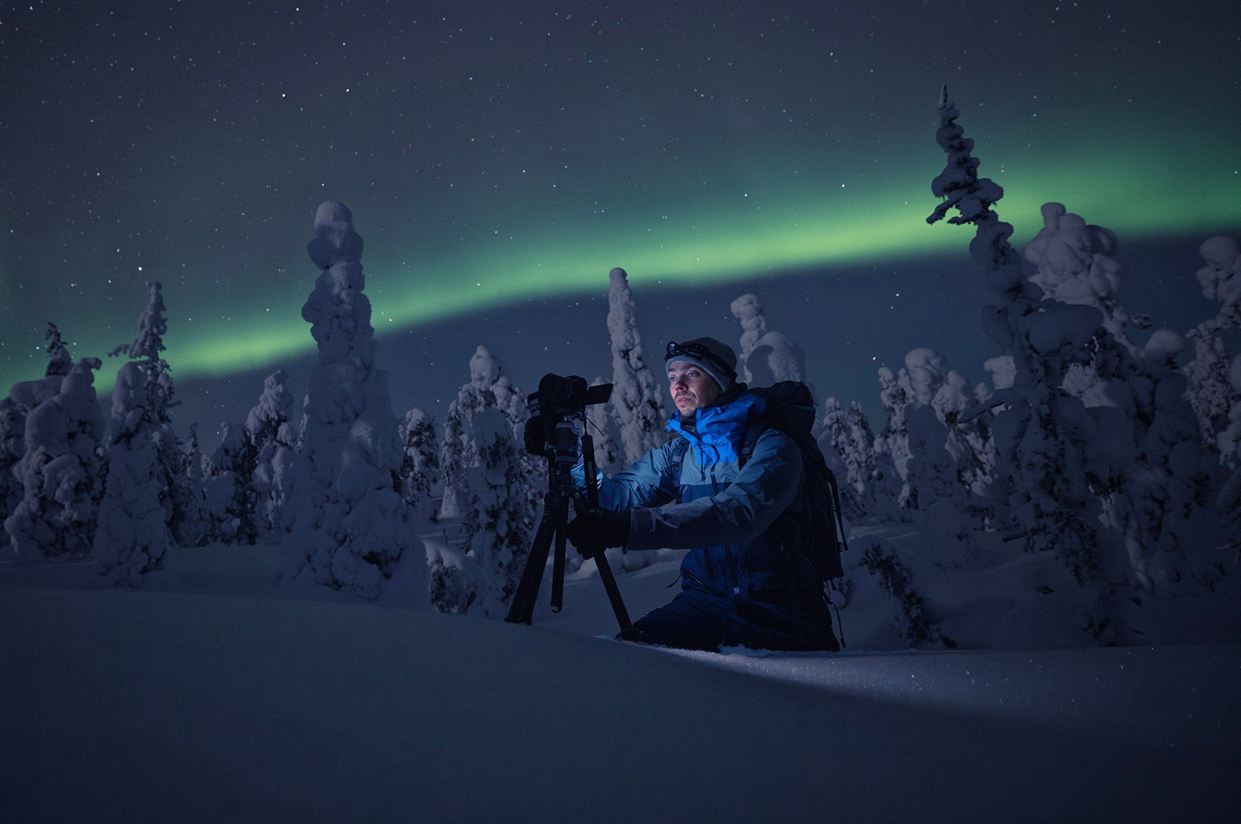 A man is kneeling down in the snow taking a picture of the aurora borealis.