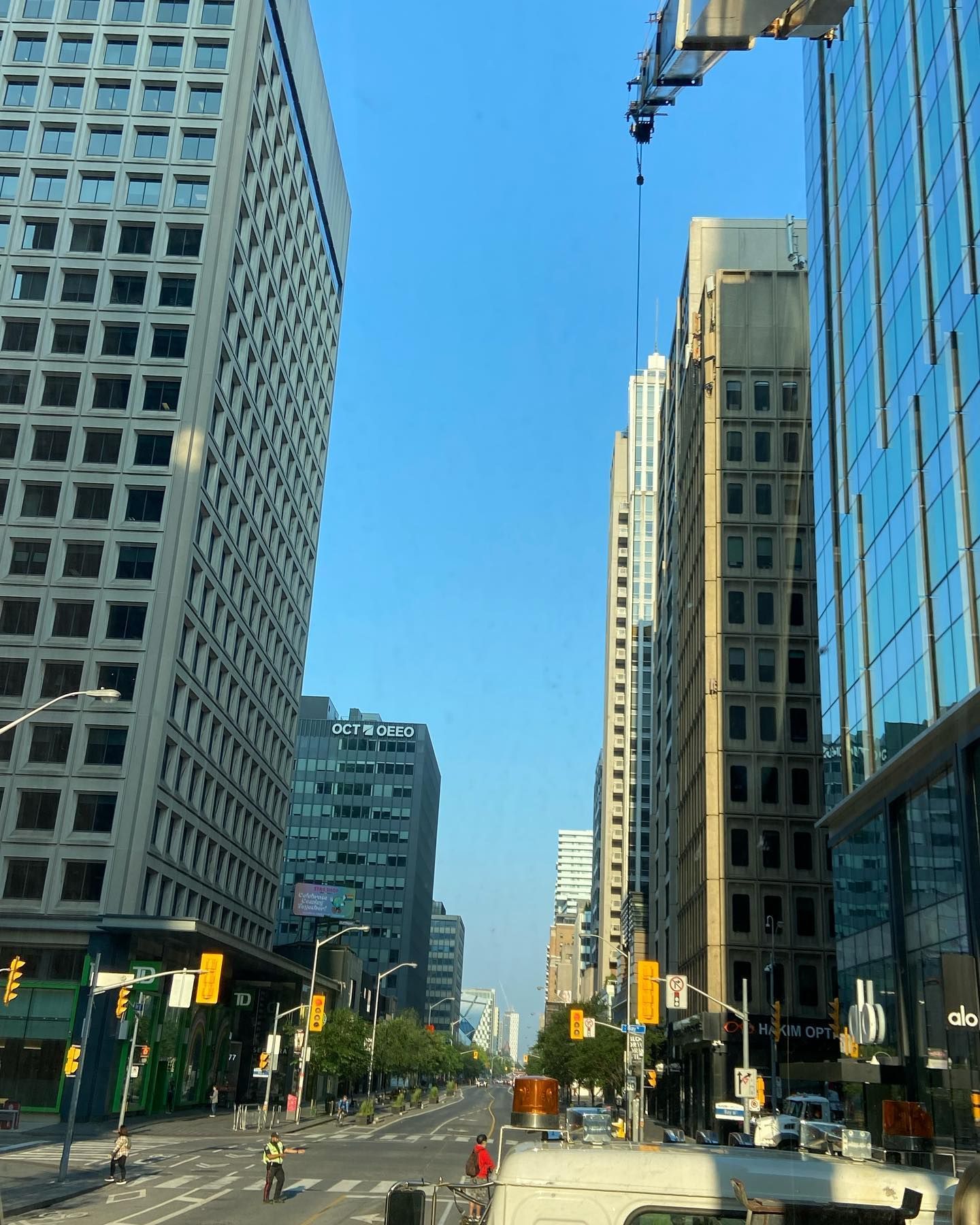 A city street intersection viewed from a vehicle, featuring tall skyscrapers, traffic lights, and a hanging crane.